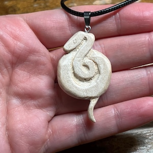 May include: A light-colored, carved wooden pendant in the shape of a coiled snake. The pendant is suspended from a black cord necklace with a silver-colored clasp. The snake's details are visible, including its head and tail.