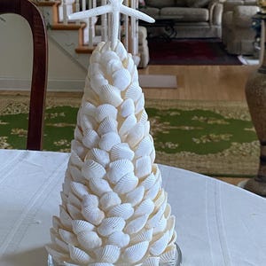 May include: A decorative, cone-shaped tree made of white seashells, topped with a white starfish. The tree sits on a clear glass plate and is displayed on a white tablecloth. The tree is a coastal or nautical themed item.