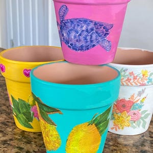 May include: A collection of hand-painted terracotta flower pots. The pots are painted in bright colors, including pink, yellow, and turquoise. Designs include a turtle, lemons, and floral patterns. Each pot has a unique design.