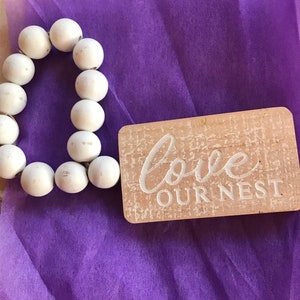 May include: A white beaded heart-shaped garland with a wooden sign that reads "love our nest" on a purple background.