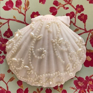 May include: A white seashell decorated with a pearl and leaf design. The shell is on a green background with a floral pattern.