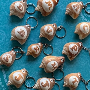 May include: A collection of 12 seashell keychains, each with a different letter of the alphabet engraved on the front. The seashells are a light brown color with a white interior. The keychains are attached to silver metal rings.