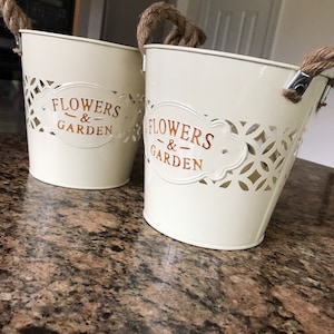 May include: Two white metal flower pots with rope handles. The pots have a cut-out design and the words "Flowers & Garden" in gold lettering.
