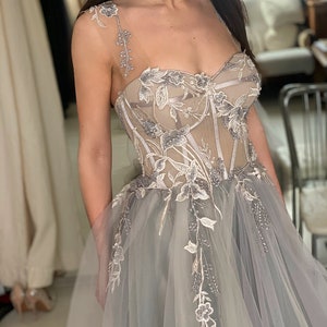 May include: A grey tulle wedding dress with floral embroidery and a fitted bodice. The dress has a sweetheart neckline and a full skirt.