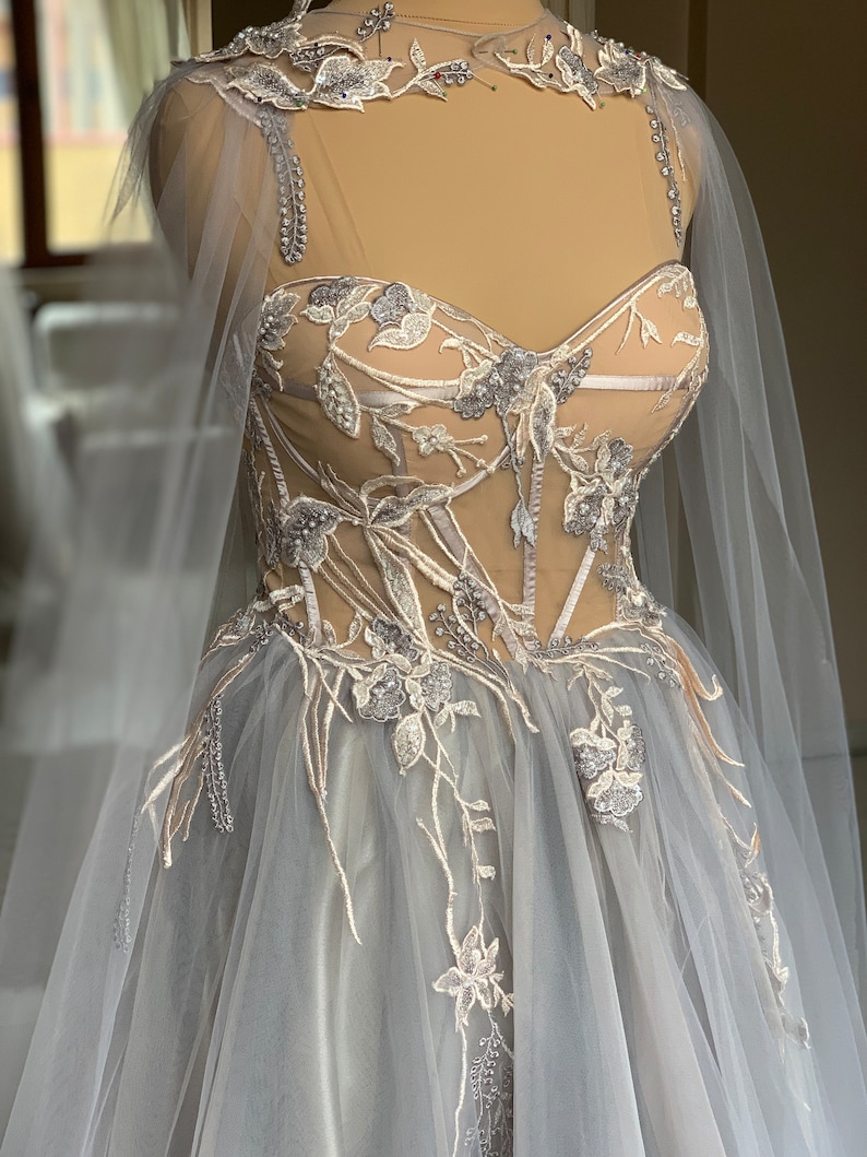 May include: A grey tulle wedding dress with a lace bodice and a cape. The bodice is decorated with floral embroidery and beads.