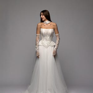 May include: A white wedding dress with a strapless, lace-detailed bodice and a flowing tulle skirt. The dress includes matching lace opera gloves and a choker. The model is standing against a grey backdrop.