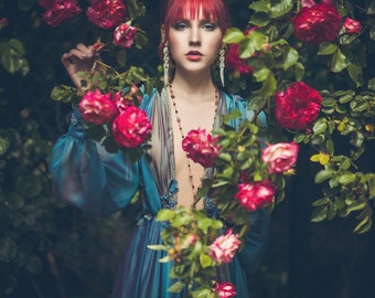 May include: A woman with bright red hair wears a teal blue silk robe with a plunging neckline. She stands in front of a bush of red roses.
