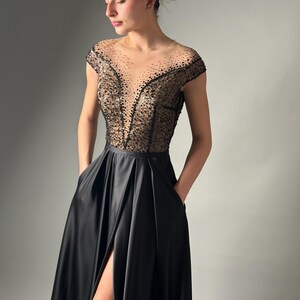 May include: A black satin gown with a beaded bodice and a high slit. The gown has a scoop neckline and cap sleeves.