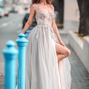 May include: A woman wearing a white, tulle gown with lace detailing and a high slit. She is standing on a pavement with blue railings in the foreground.