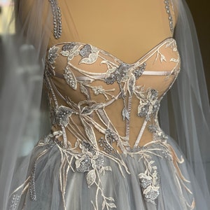 May include: A grey tulle ball gown with a fitted bodice featuring delicate floral embroidery and silver embellishments. The bodice has a sweetheart neckline and a sheer overlay. The skirt is full and flowing.
