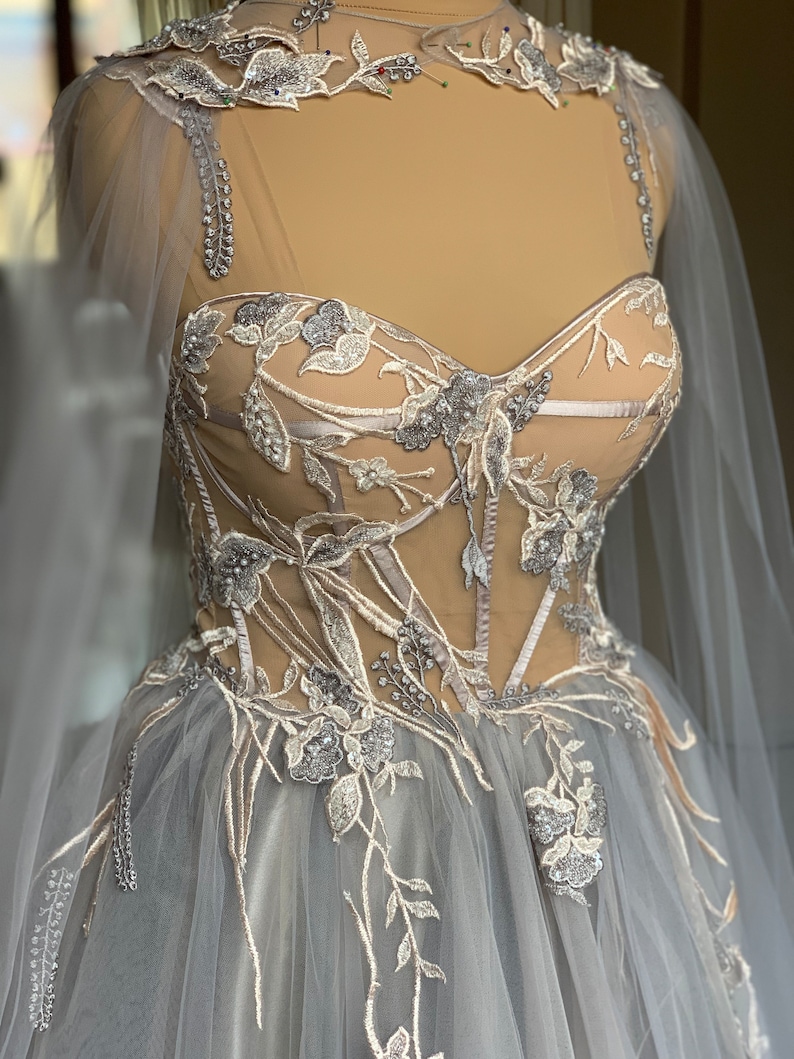 May include: A grey tulle ball gown with a fitted bodice featuring delicate floral embroidery and silver embellishments. The bodice has a sweetheart neckline and a sheer overlay. The skirt is full and flowing.