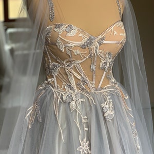 Op de afbeelding: Een grijze tule bruidsjurk met een kantcorset en een cape. Het corset is versierd met bloemenborduurwerk en kralen.