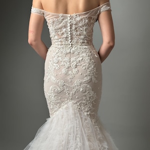 Peut inclure: Une robe de mariée blanche à épaules dénudées avec un corsage ajusté et une jupe ample. Le corsage est recouvert de dentelle et de perles complexes, et la jupe est faite de plusieurs couches de tulle.