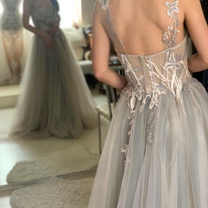 May include: A light grey tulle wedding dress with delicate floral embroidery and a fitted bodice. The dress has a flowing skirt and a low back.