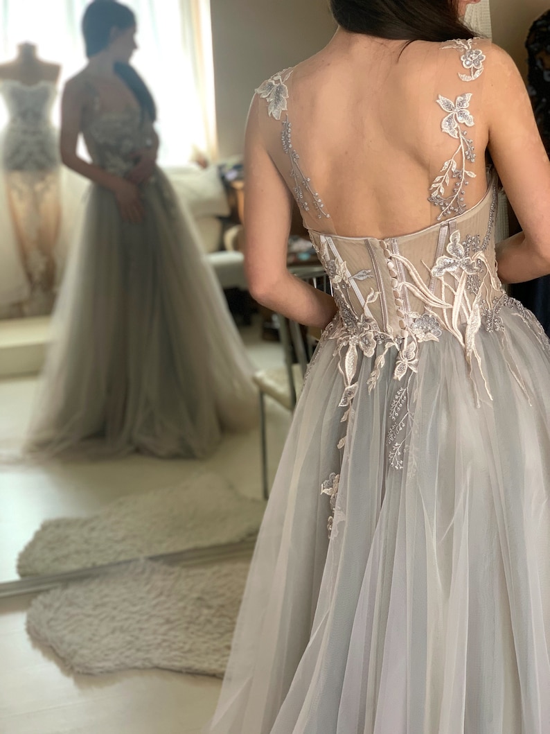 May include: A light grey tulle wedding dress with delicate floral embroidery and a fitted bodice. The dress has a flowing skirt and a low back.