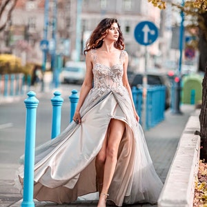 May include: A woman in a long, flowing, light-coloured dress with a high slit walks down a city street. The dress has a lace bodice and a flowing skirt. The woman is wearing silver sandals.