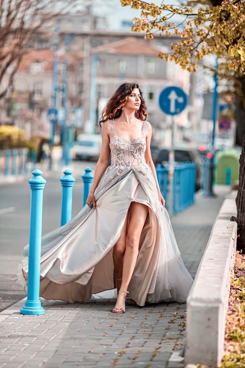 May include: A woman in a long, flowing, light-coloured dress with a high slit walks down a city street. The dress has a lace bodice and a flowing skirt. The woman is wearing silver sandals.