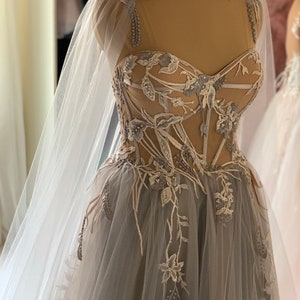 May include: A grey tulle gown with a sheer bodice and floral embroidery. The gown has a long train and a matching cape.
