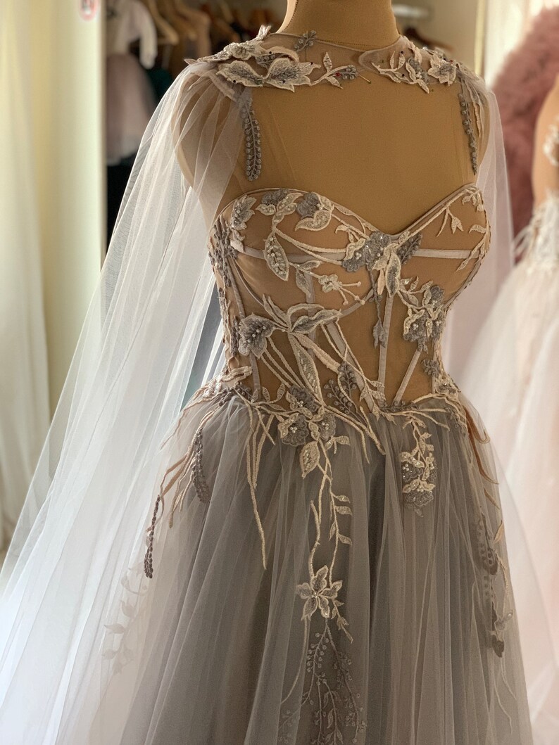 May include: A grey tulle gown with a sheer bodice and floral embroidery. The gown has a long train and a matching cape.