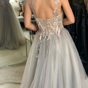 May include: A light grey tulle gown with delicate floral embroidery and a sheer bodice. The gown has a fitted bodice and a flowing skirt.