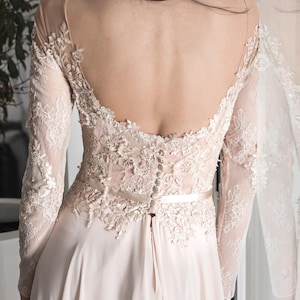 Puede incluir: Un vestido de novia de encaje blanco con la espalda descubierta y mangas largas. El vestido tiene un corpiño ajustado y una falda fluida.