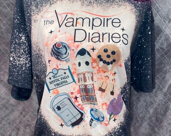 Puede incluir: Camiseta gris oscuro con un diseño blanco blanqueado con el texto "The Vampire Diaries" y varias ilustraciones. La camiseta incluye imágenes de edificios, comida y las palabras "Welcome to Mystic Falls Virginia."