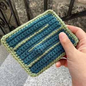 May include: A small crocheted square with a teal blue background and green stripes. The square is made with a single crochet stitch.