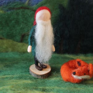 Könnte beinhalten: Eine handgefertigte Filz-Wichtelfigur mit langem weißem Bart, roter Mütze und grünem Mantel, stehend auf einem Holzsockel. Daneben liegt ein aufgerollter orangefarbener Filzfuchs. Die Szene vor grünem und dunklem Hintergrund.