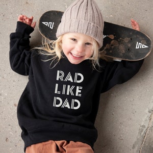 以下が含まれることがあります： 白い文字で「RAD LIKE DAD」と書かれた黒いスウェットシャツを着た子供。子供は薄いピンクのビーニーをかぶり、スケートボードを持っています。