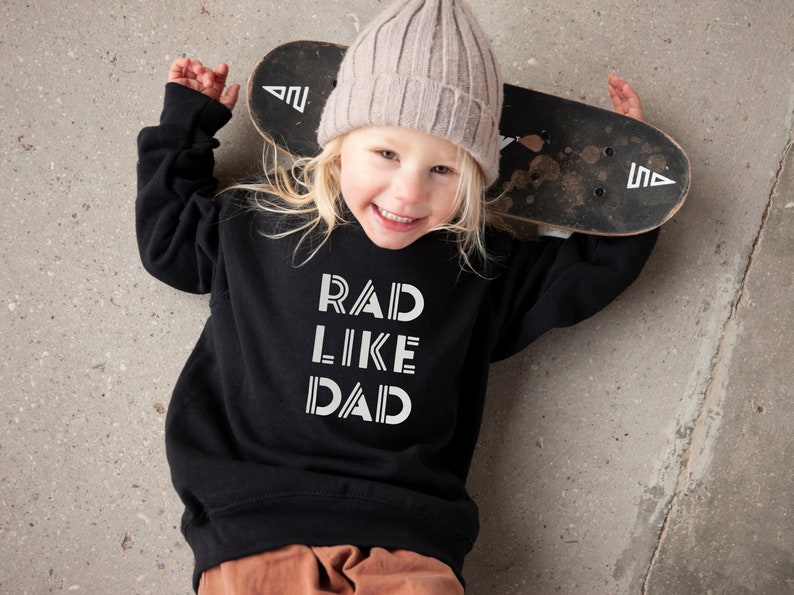 以下が含まれることがあります： 白い文字で「RAD LIKE DAD」と書かれた黒いスウェットシャツを着た子供。子供は薄いピンクのビーニーをかぶり、スケートボードを持っています。