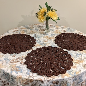 May include: Three brown crocheted placemats on a white tablecloth with a floral pattern. A vase with yellow flowers sits in the center of the table.