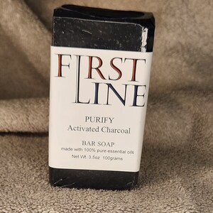May include: A black bar of soap with a white label. The label reads "FIRST LINE" in red and blue lettering, with the words "PURIFY Activated Charcoal BAR SOAP" below. The net weight is 3.5oz (100 grams).