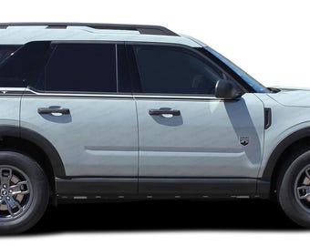 May include: A light gray Ford Bronco Sport SUV with black trim and black wheels. The vehicle has a black roof rack and the word "SPORT" is visible on the side of the vehicle.