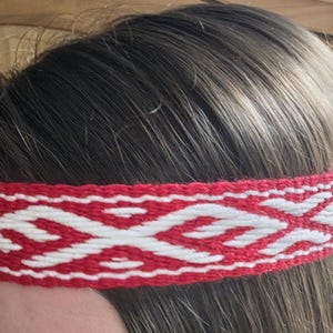 May include: A red and white woven headband with a geometric pattern. The headband is wrapped around a person's head, with the ends hanging down. The design features a repeating pattern of white shapes on a red background.