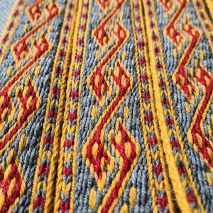 May include: Close-up of a woven fabric with a repeating pattern of red, yellow, and gray diamonds. The fabric has a textured surface and a slightly blurred background.