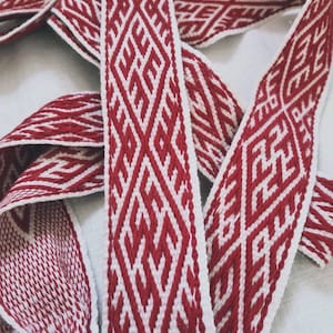 May include: Woven red and white belt with a geometric diamond pattern. The belt is made of a sturdy material and features a repeating design. The edges are finished with a white border, and the belt is folded over itself.