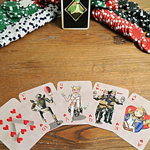 May include: A set of playing cards featuring characters from the video game Apex Legends. The cards are arranged in a semi-circle on a wooden table. The cards are all hearts and include a 10, Jack, Queen, King, and Ace.