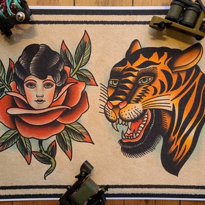 Op de afbeelding: Een vintage tattoo-flash sheet met twee ontwerpen. Het eerste ontwerp is een vrouwengezicht in een rode roos. Het tweede ontwerp is een grommend tijgerhoofd met oranje en zwarte strepen.