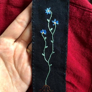 Peut inclure: Un marque-page en tissu noir avec un motif brodé de fleurs bleues avec des tiges vertes et des racines brunes.