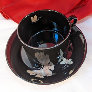 Puede incluir: Una taza de té y un platillo negros con un delicado diseño de mariposa. La taza y el platillo están decorados con mariposas y libélulas plateadas.