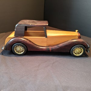 May include: A wooden toy car in a classic style. The car has a natural wood body with dark brown accents, gold-coloured wheels, and a dark brown roof. The toy car is a vintage-style collectible.