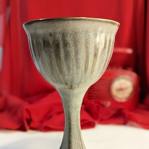 Puede incluir: Un cáliz de cerámica gris con un diseño acanalado. El cáliz tiene un tallo alto y delgado y una base ancha y redonda.