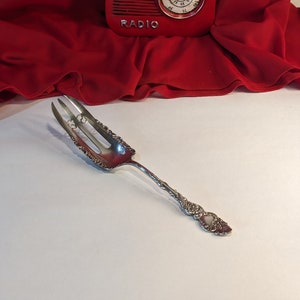 May include: A silver serving fork with intricate floral patterns. The fork features three prongs and a decorative handle. Behind it, a red retro radio with the word "RADIO" is set against a red fabric background.