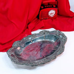 May include: A silver-colored, oval-shaped serving tray with ornate, scrolling edges. The tray has a slightly tarnished appearance, suggesting age or use. A red vintage radio is visible in the background, along with a red fabric.
