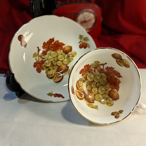 Puede incluir: Una taza de té y un platillo blancos con un borde dorado. El diseño presenta un racimo de uvas verdes, nueces marrones y hojas rojas.
