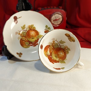 Puede incluir: Una taza de té y un platillo blancos con un borde dorado. La taza de té y el platillo presentan un diseño de frutas coloridas con manzanas, bayas y hojas.