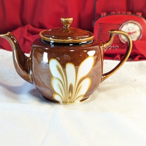 Peut inclure: Une théière en céramique brune avec un motif floral blanc. La théière a un couvercle et une poignée.
