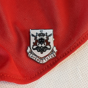Op de afbeelding: Een zilveren en witte emaille pin met een wapenschildontwerp. Het schild heeft een zwart en bruin ontwerp met twee witte herten. De tekst "CAVENDO TUTUS" staat op een rode banner onder het schild.