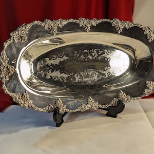 May include: A silver oval serving tray with a grapevine design around the edge. The tray has a raised rim and a smooth, reflective surface.