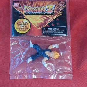 Peut inclure: Un sac en plastique contenant une petite figurine d'action aux cheveux jaunes d'un personnage de la série d'anime Dragonball Z. La figurine est allongée sur le dos, les bras tendus. Le sac contient également une carte avec le logo Dragonball Z et le texte "The Saga Continues". La carte comprend également le texte "Item #3845" et "Ages 4 & up".